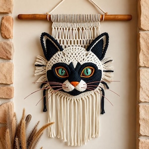 Peut inclure: Une suspension murale en macramé représentant un visage de chat stylisé en corde crème et noire. Le chat a des yeux vert vif, un nez orange et de longues moustaches fines. L'ensemble est suspendu à une tige en bois avec une corde crème.