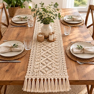 Peut inclure: Un chemin de table en macramé couleur crème présentant un motif géométrique tissé complexe et de longues franges aux extrémités. Le chemin est centré sur une table à manger en bois avec des assiettes blanches, des serviettes et des verres.
