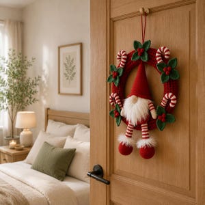 May include: A crocheted Christmas wreath featuring a central gnome with a red hat, white beard, and striped legs. The wreath is decorated with green crocheted holly leaves, red berries, and red and white striped candy canes hanging on a door.