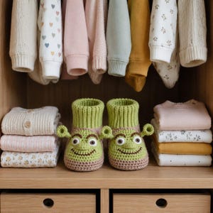Peut inclure: Une paire de chaussons en crochet vert clair représentant un visage de personnage avec des oreilles en forme de trompette, de grands yeux et un large sourire. Ils ont un revers côtelé et une base beige, posés sur une étagère en bois.