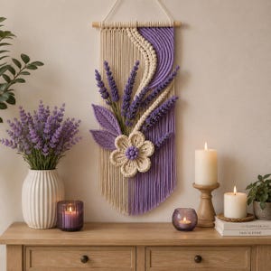 May include: A macrame wall hanging featuring cream and purple cotton cords, a large cream flower with a purple center, and lavender sprigs. The piece is suspended from a wooden dowel with a white cord, showcasing intricate knotting and fringe.