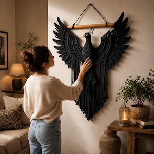 May include: A black macrame bird wall hanging with outstretched wings, suspended from a wooden dowel. The bird features intricate detailing and a textured design. This decorative wall art is a stylish home decor item.