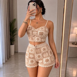Peut inclure: Un ensemble deux pièces en crochet composé d'un haut court à bretelles fines et d'un short taille haute. Le vêtement présente un motif répétitif de carrés granny dans des tons beige, crème et brun clair, avec un ourlet festonné sur le short.