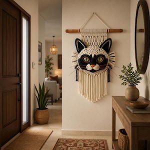 May include: A macrame wall hanging featuring a stylized cat face with cream and black knotted cord, green eyes, and a small orange nose. The piece is suspended from a wooden dowel with long, cream-colored fringe hanging from the bottom edge.
