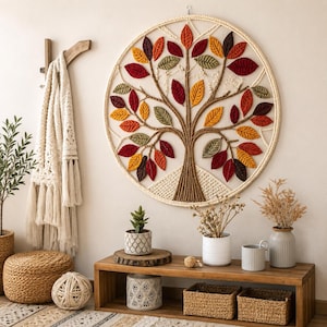 Peut inclure: Une décoration murale circulaire représentant un arbre en macramé avec des feuilles en tissu dans des tons de rouge, orange, jaune et vert. Le tronc et les branches sont faits de corde torsadée, fixés dans un cadre circulaire crème.