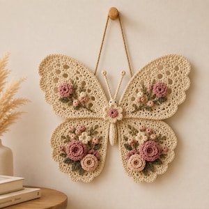 Op de afbeelding: Een crèmekleurige gehaakte vlinder als wanddecoratie met opengewerkte vleugels, versierd met driedimensionale roze en witte bloemclusters en groene blaadjes. De vlinder hangt aan een dun bruin koord aan een kleine houten knop.