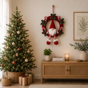 May include: A red crocheted Christmas wreath featuring a central gnome figure with a tall pointed hat, a white beard, and striped legs. The wreath is decorated with green fabric leaves and small red and white striped candy cane ornaments.