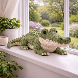 Op de afbeelding: Een handgemaakt gehaakt alligatorspeelgoed in groen- en crèmetinten, met zwarte knoopogen. De alligator ligt op een witte vensterbank, met een tuin zichtbaar door het raam. Het pluche speelgoed is een decoratief item.