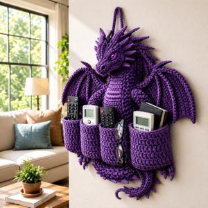 May include: A handcrafted, purple crochet dragon wall organizer with four pockets. The dragon has wings, a tail, and a head with two purple eyes. The pockets hold remotes, glasses, and small books. The organizer is designed to hang on a wall.