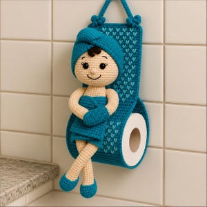 May include: A crocheted toilet paper holder featuring a doll with beige skin, dark hair, and a teal towel wrap on its head and body. The holder has a teal backing with a small heart pattern and a cylindrical compartment for a roll of toilet paper.