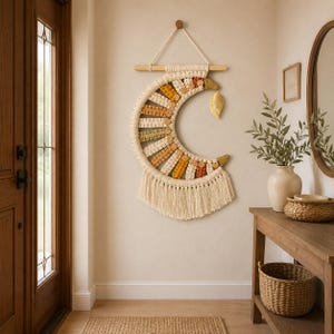 May include: A crescent moon-shaped macrame wall hanging made of off-white cord, featuring alternating sections of mustard yellow, burnt orange, and sage green yarn. The piece includes wooden beads, a gold-coloured leaf charm, and long fringe.