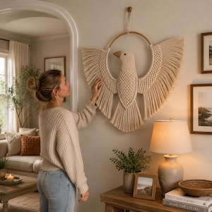 May include: A beige macrame wall hanging in the shape of a bird with outstretched wings. The bird is centered within a wooden hoop. The wings and tail feature long, fringed edges. The bird's body is intricately knotted, and it has a wooden beak.