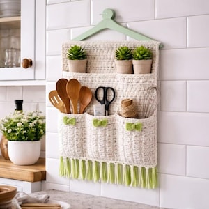Op de afbeelding: Een crèmekleurige, gehaakte keukenorganizer met drie zakken en twee planken, hangend aan een groene hanger. De zakken zijn versierd met groene strikken en franjes. Het bevat houten lepels, een schaar en kleine potplanten.