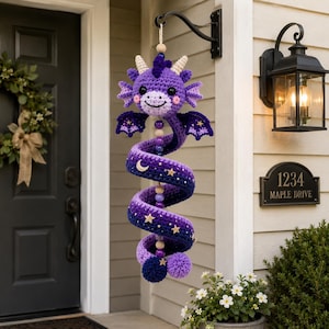 May include: A purple crochet dragon wind spinner featuring a spiral body with an ombre effect from light to dark purple. The dragon has small wings, horns, and a smiling face. The spiral is decorated with small wooden beads, stars, and a moon.