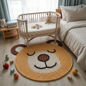 Peut inclure: Un tapis rond en crochet en forme d'ours, aux tons jaune moutarde, crème et marron foncé. Le tapis représente un visage d'ours avec des yeux fermés, un nez marron foncé et deux oreilles arrondies. Il est posé sur un sol en bois clair.