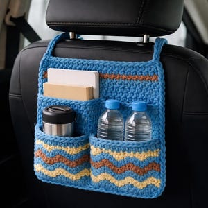 Peut inclure: Organisateur de voiture en crochet bleu avec plusieurs poches. Il se suspend à l'appuie-tête du siège auto et contient des bouteilles d'eau, un thermos et des cahiers. Les poches présentent un motif en zigzag jaune et marron.