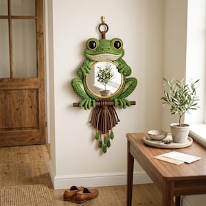 May include: A wall-mounted mirror featuring a green crocheted frog frame with large black eyes. The frog sits on a horizontal wooden dowel, with a brown macrame fringe hanging below, accented by small green beads. The mirror is circular with a gold rim.