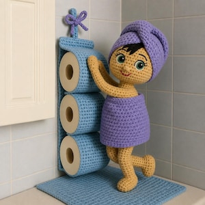 Peut inclure: Un porte-papier toilette en crochet bleu clair avec une boucle de suspension et trois compartiments cylindriques. À côté se trouve une poupée en crochet au teint hâlé, cheveux noirs, portant une serviette violette sur la tête et le corps.