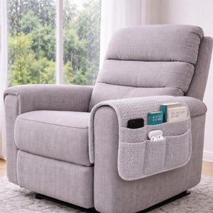 May include: A light gray upholstered recliner chair with a built-in side pocket organizer. The organizer holds a black phone, white earbuds, and two small books. The chair is positioned in front of a window.