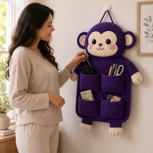 Könnte beinhalten: Ein lila gehäkelter, affenförmiger Wandorganizer mit mehreren Taschen. Der Affe hat ein cremefarbenes Gesicht, Ohren und Pfoten. Der Organizer hängt an einer weißen Wand und ist mit verschiedenen Gegenständen gefüllt.