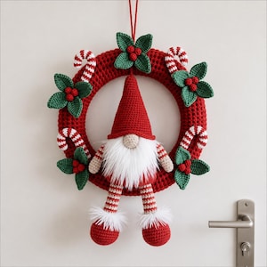 May include: A crocheted Christmas wreath featuring a red circular base adorned with green holly leaves, red berries, and red and white candy canes. A gnome with a red hat, white beard, and striped legs hangs from the center of the wreath.