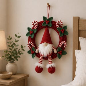 May include: A circular crochet wreath in deep red featuring a central gnome with a tall red hat, white beard, and dangling striped legs. The wreath is decorated with crocheted green holly leaves, red berries, and white and red candy canes.