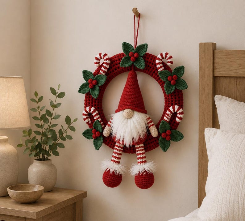 May include: A circular crochet wreath in deep red featuring a central gnome with a tall red hat, white beard, and dangling striped legs. The wreath is decorated with crocheted green holly leaves, red berries, and white and red candy canes.