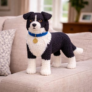 Peut inclure: Figurine de chien crocheté noir et blanc, avec un collier bleu et une médaille dorée. Le chien a une texture duveteuse et est posé debout sur une surface beige. Les yeux du chien sont bruns.