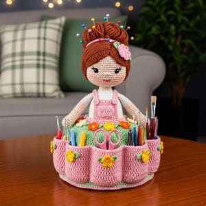 Peut inclure: Une poupée au crochet faite à la main avec un chignon brun orné d'épingles et d'une fleur rose. La poupée porte une robe rose et tient un récipient avec des poches pour les fournitures d'artisanat. Le récipient est rose avec des accents de fleurs jaunes.