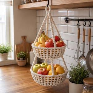 Op de afbeelding: Een cr&egrave;mekleurige macram&eacute; fruitmand met twee niveaus, gevuld met vers fruit, waaronder rode en groene appels, bananen en citroenen. De mand hangt aan een zwarte haak en voegt een vleugje bohemienstijl toe aan de keuken.