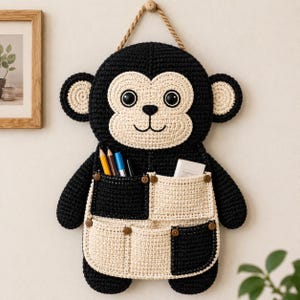 Könnte beinhalten: Ein gehäkelter, affenförmiger Wandorganizer in Schwarz und Creme mit einer Aufhängeschnur. Der Affe hat ein cremefarbenes Gesicht, Ohren und Bauch mit schwarzen Akzenten. Er hat zwei Taschen mit Holzknöpfen, in denen Stifte und kleine Gegenstände aufbewahrt werden.