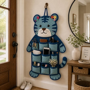 May include: A blue and light blue crochet tiger-shaped wall organizer with multiple pockets. The tiger has a white face, brown nose, and colorful button accents. The organizer is hanging on a wall and holds keys, a phone, and other small items.
