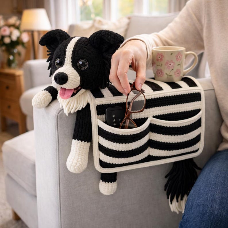 Puede incluir: Un peluche de ganchillo de un border collie en blanco y negro unido a un organizador de reposabrazos de tela a rayas blancas y negras con dos bolsillos. El organizador contiene unas gafas de montura marr&oacute;n y una taza con flores rosas.