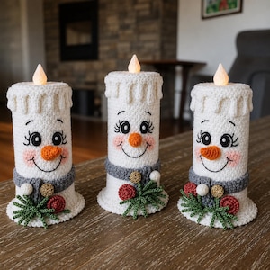 Peut inclure: Trois housses de bougie en crochet blanc représentant des visages de bonhomme de neige avec des nez orange, des yeux noirs et des joues roses. Chacune porte une écharpe grise, des brins de pin verts, des baies rouges et un détail doré.
