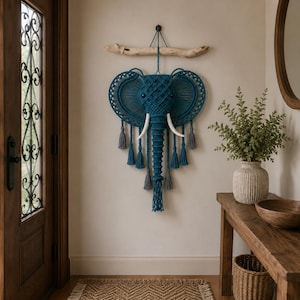 Peut inclure: Décoration murale en macramé représentant un éléphant bleu sarcelle avec des défenses blanches et des pompons gris. L'éléphant est suspendu à une branche de bois flotté. Décoration murale.