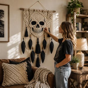 May include: A macrame wall hanging featuring a skull design. The skull is cream-colored with black eye sockets and teeth. Black tassels and feather accents hang below. The wall hanging is suspended from a wooden dowel.