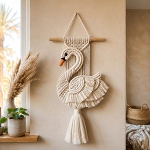 Peut inclure: Une suspension murale en macramé couleur crème en forme de cygne, accrochée à une tige en bois horizontale. Le cygne présente des motifs noués complexes, un œil noir et un bec orange, avec un long pompon à franges suspendu en bas.