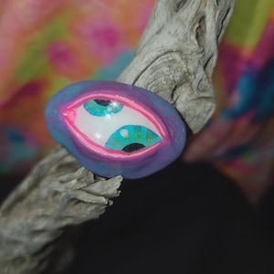 May include: A hand-painted, oval-shaped art piece featuring two stylized eyes. The eyes have turquoise irises, black pupils, and are outlined in pink and purple. The artwork is set against a backdrop of a weathered wooden branch and a colorful, abstract background.