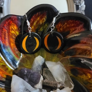 May include: A pair of black and orange crescent moon earrings with silver hooks. The earrings are displayed on a colorful surface with crystals.