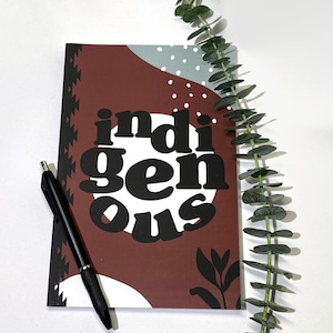 May include: A brown notebook with a white and gray geometric design. The notebook has the word "indigenous" written in black letters on the front cover. A black pen is lying on the notebook.