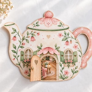 Può includere: Una sagoma in legno piatta a forma di teiera con un motivo floreale dipinto nei toni del rosa e del verde. La teiera include una piccola porta in legno incernierata che si apre per rivelare una scena interna stampata di una stanza.