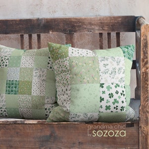 以下が含まれることがあります： 木製のベンチの上に、緑と白の組み合わせのパッチワークのクッションが2つ置かれています。クッションには花柄とクローバー柄が施され、"grandma chic by sozoza"というラベルが付いています。