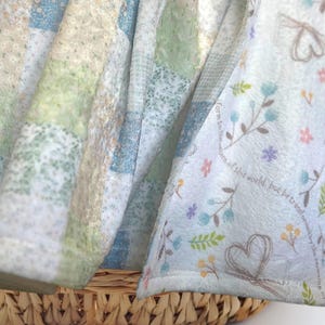 May include: A soft, white blanket with a patchwork design in shades of green, blue, and beige. The blanket has a floral print with the text "form to us...warm of this world, but be transformed..."