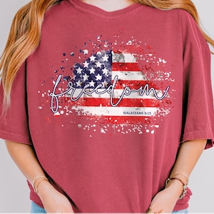 Può includere: Una maglietta bordeaux con un disegno della bandiera americana invecchiata e la parola "freedom" in corsivo. Il testo "GALATIANS 5:13" è stampato sotto. La maglietta ha maniche corte e girocollo.