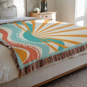 May include: A woven throw blanket with a retro sunburst design in shades of orange, yellow, teal, and red. The blanket features the text "His mercies are new every morning. Great is His faithfulness."