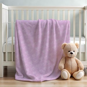 Könnte beinhalten: Eine weiche, plüschige Lavendeldecke mit einem weißen, sich wiederholenden Muster des Wortes "Little" darauf. Ein hellbrauner Teddybär sitzt auf der Decke.