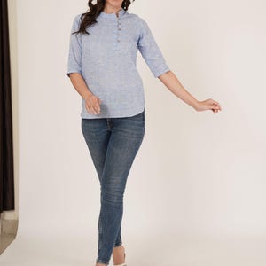 May include: A woman wearing a light blue, short-sleeved, button-up top with a white collar and blue jeans. She is wearing white sandals.