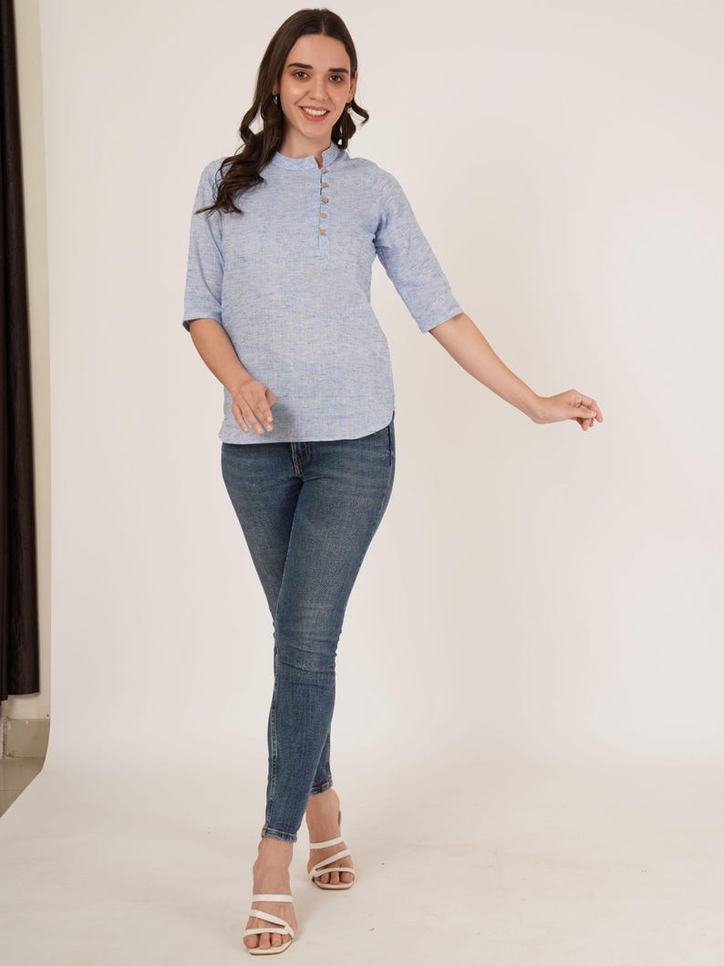 May include: A woman wearing a light blue, short-sleeved, button-up top with a white collar and blue jeans. She is wearing white sandals.