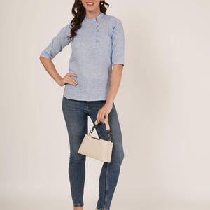 May include: A woman wearing a light blue, button-down shirt with a white handbag and jeans.