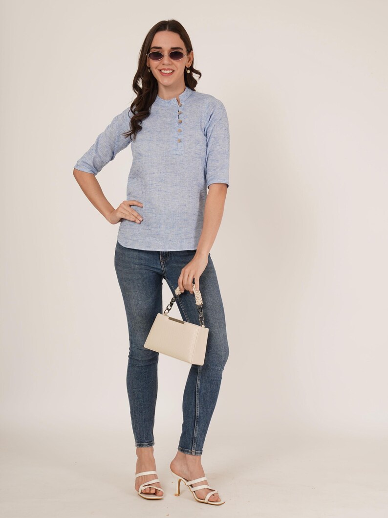 May include: A woman wearing a light blue, button-down shirt with a white handbag and jeans.